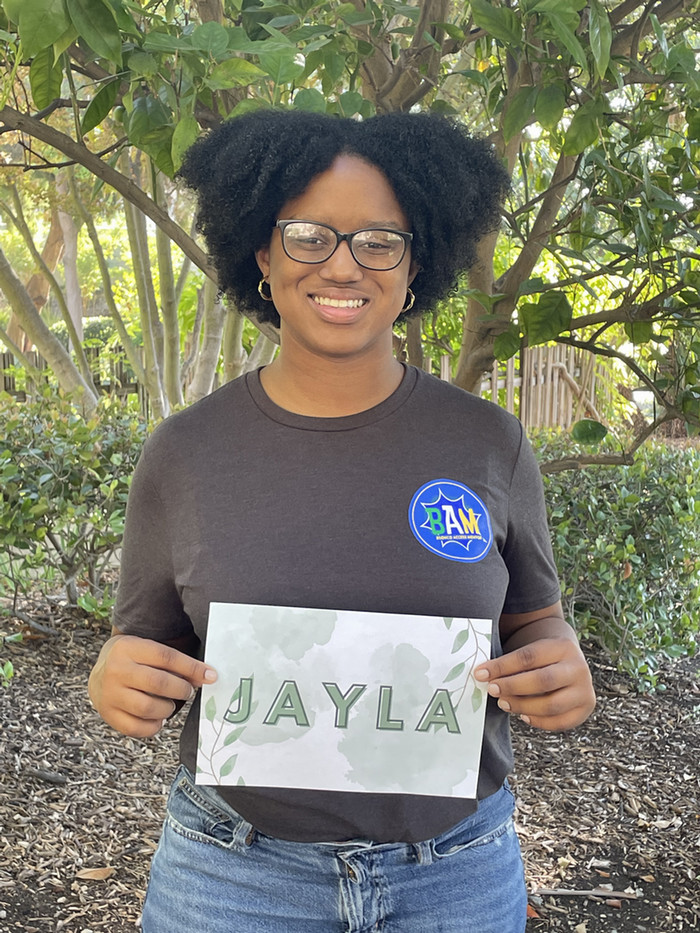 Jayla is smiling under a tree holding a sign with her name on it while wearing a shirt with the BAM logo on it.