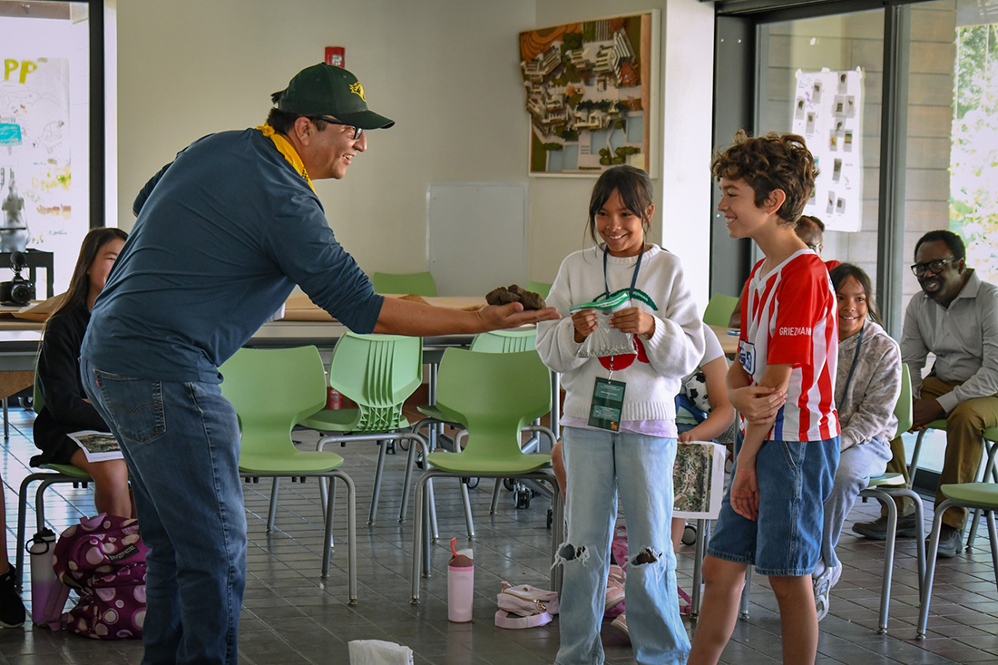 An instructor shows two students something in the palm on his hand and they are smiling. 