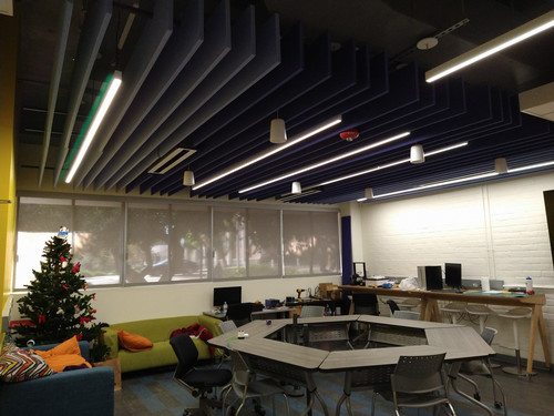 iLab social lounge area featuring a dark exposed ceiling with angled acoustic panels and linear lights above collaborative tables, seating, and a casual gathering space.