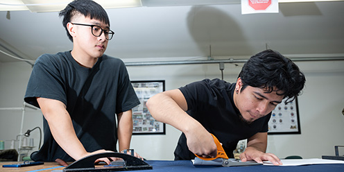 A male student cuts a garment while another looks on.