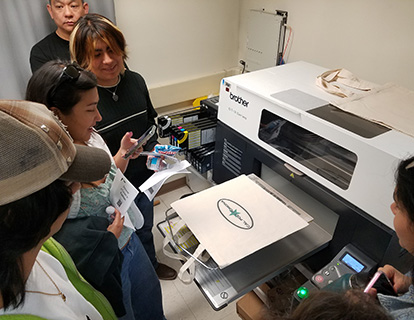 Visitors watch a printer print a label on a tote bag.