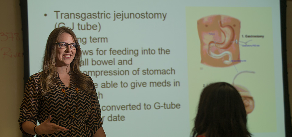 A women stands in front of a PowerPoint presentation and speaks.