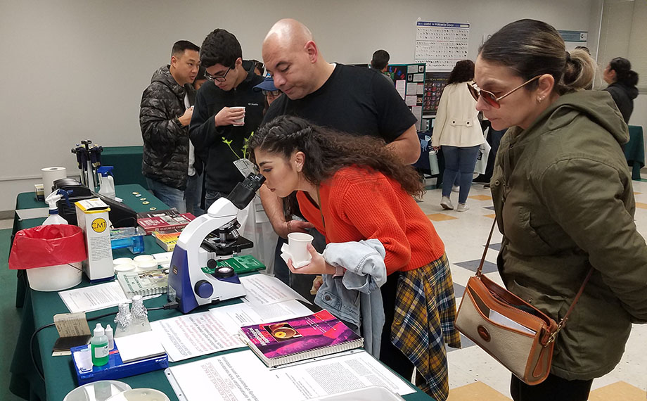 A woman peers into a microscope while a man and woman look on at open house.