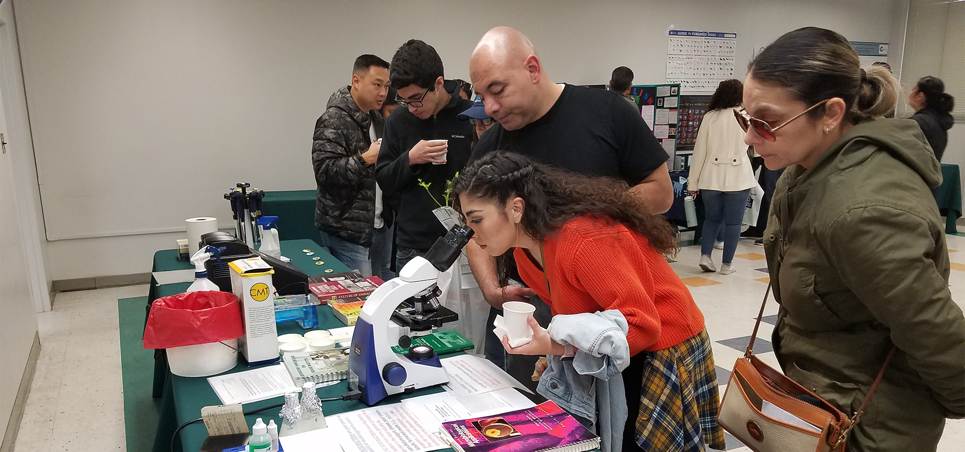 A young woman looks through a microscope at a 2020 Ag Open House display, while an older man and woman (presumably her parents) look on.