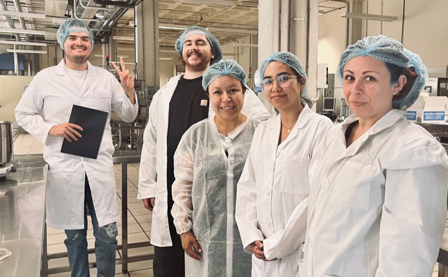 Students in lab coats and hair nets at a Spanish food processing facility.