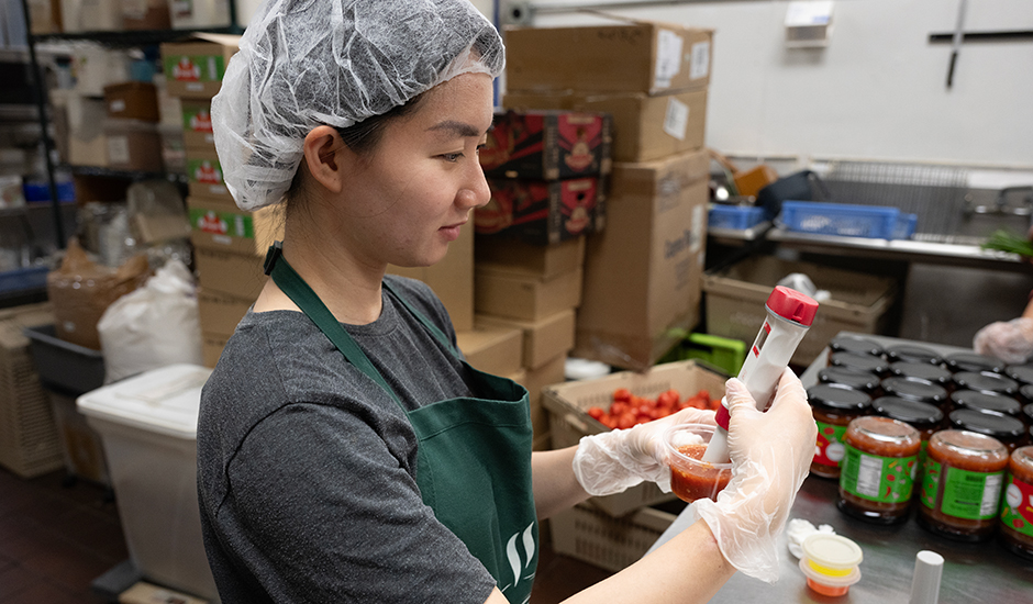 A Cal Poly Pomona student, wearing a hairnet, apron, and gloves, prepares salsa in a food production facility.