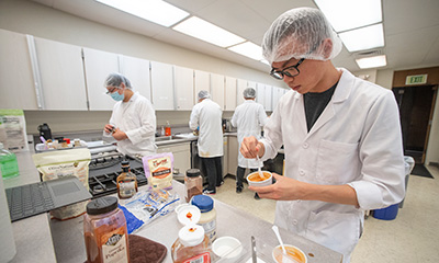 A Cal Poly Pomona food science and technology student in a hair net and white lab coat mixes food ingredients in a kitchen lab.