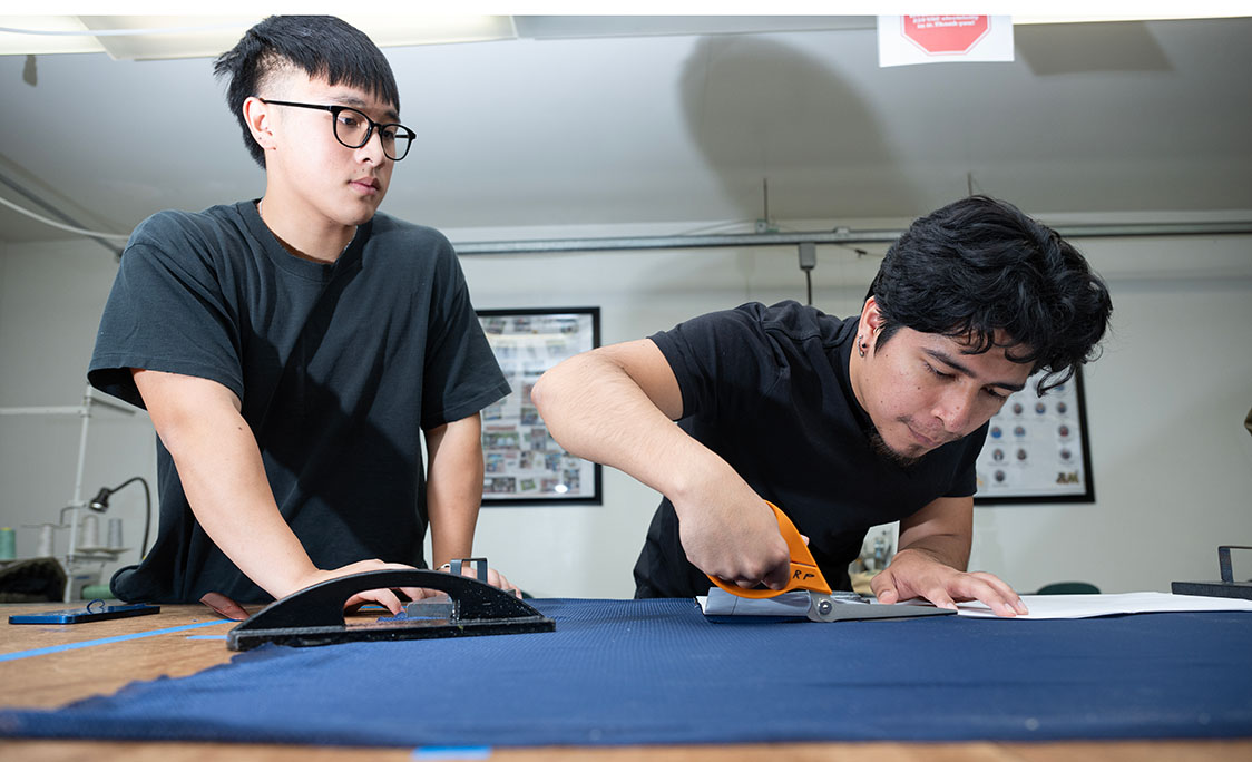A man cuts fabric on a table while another looks on.