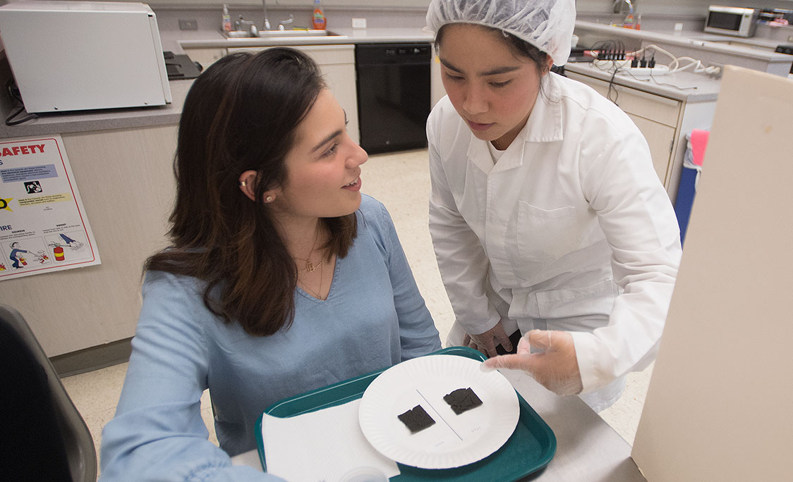 A woman in lab coat offers a food sample to another woman.