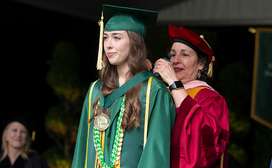 An older woman drapes a medallion around a younger woman's neck while a man watches.
