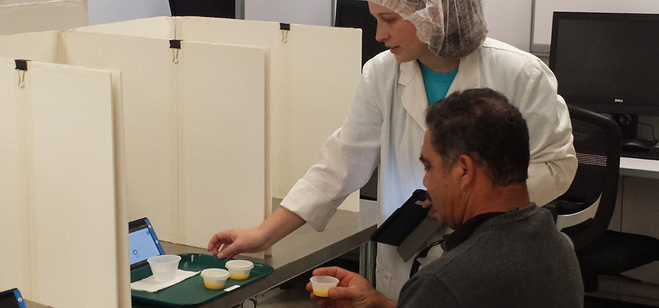 A man taste tests a small cup of orange juice as a researcher looks on.