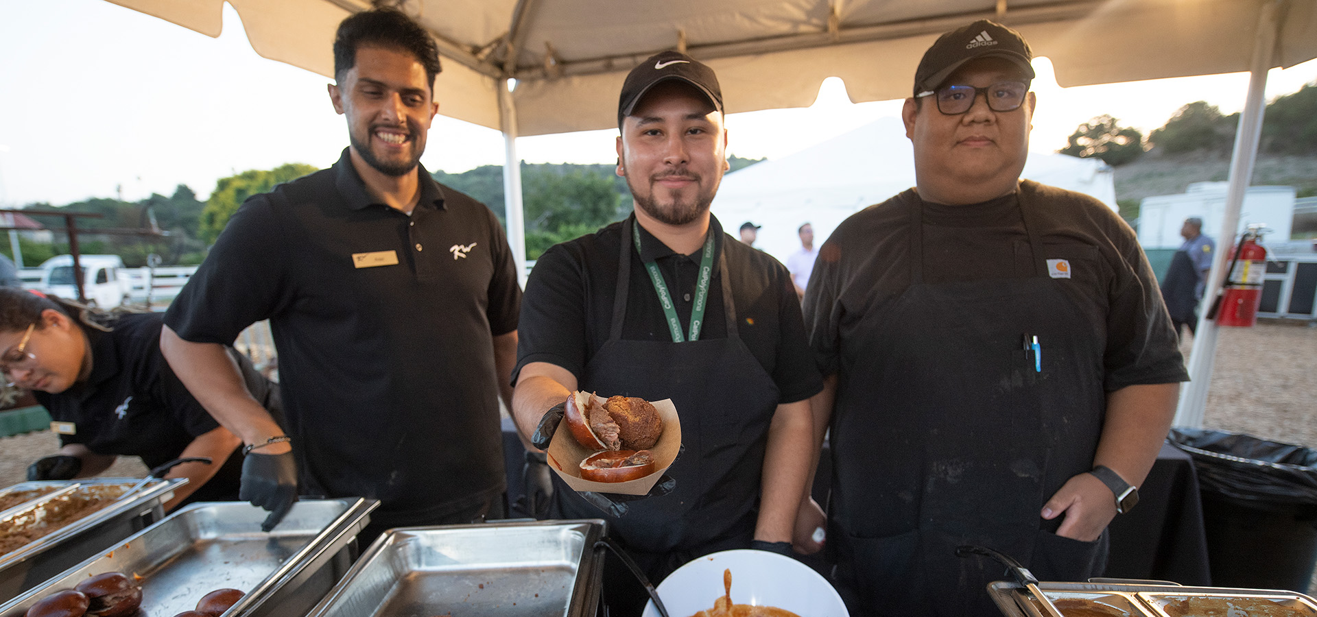 Three cooks pose for a photo at food vending tent with the center man offering up a slider in a paper or cardboard tray