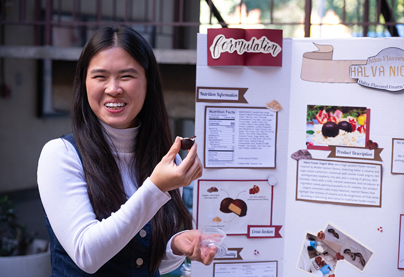 A woman smiles and holds up a food sample in front of a poster presentation.