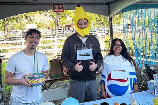 Three nutrition students pose at a booth, one has a banana-like hat on, another is dressed like a Pepsi can, and a third holds a basket of treats.