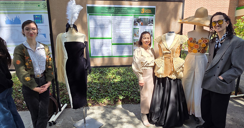 Three apparel students stand in front of their poster presentation, which includes three mannequins in dresses.