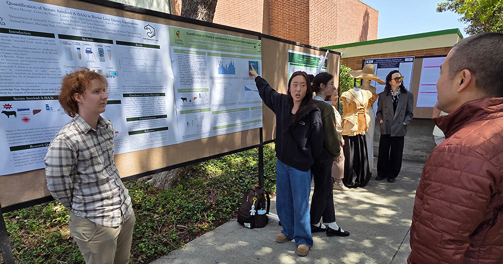 A female student raises her arm and gestures at her poster presentation, while two other students look on.