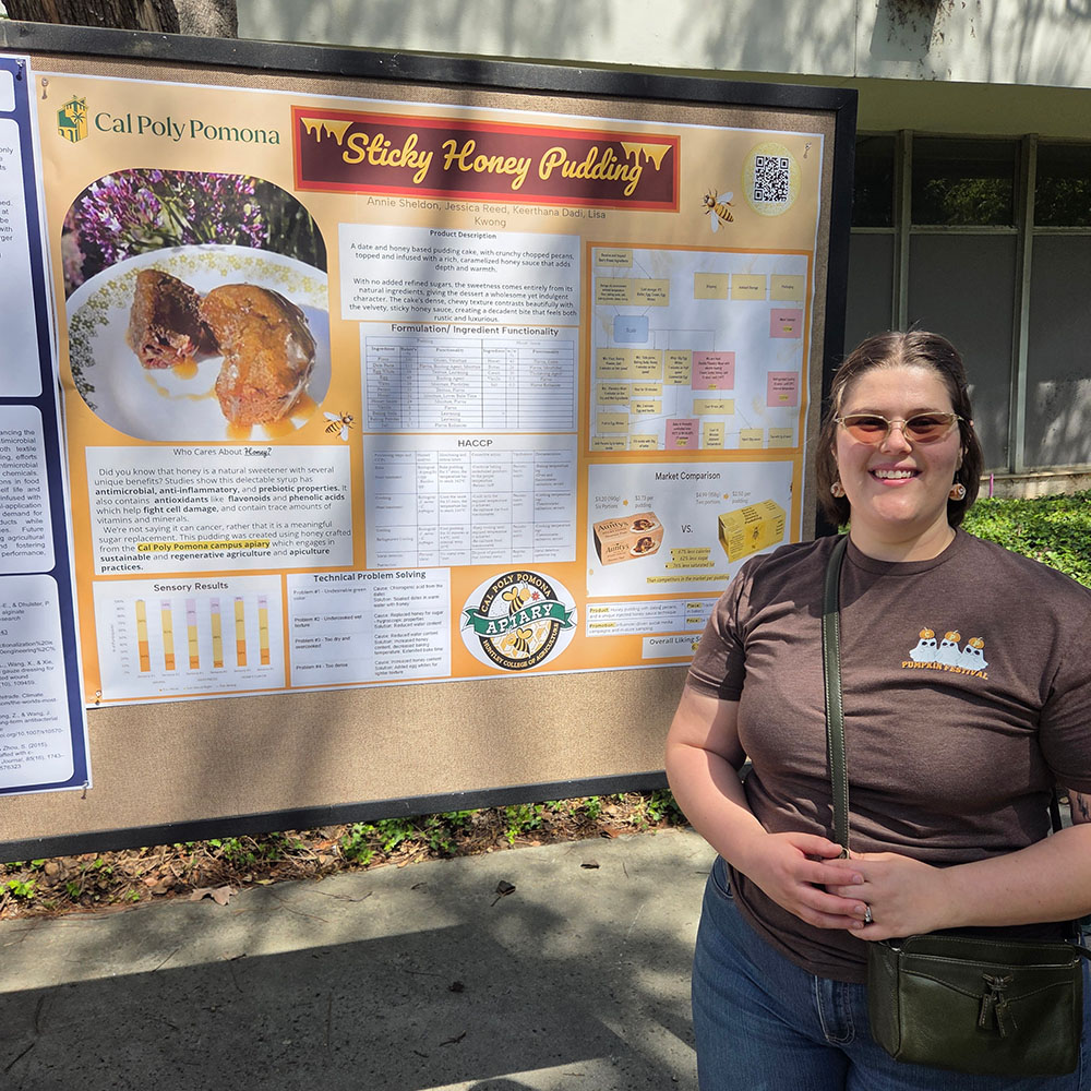 A woman in tinted glasses smiles as she stands in front of her poster presentation on "Sticky Honey Pudding."