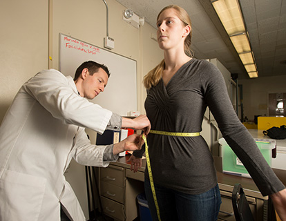 A male student in a lab coat uses a tape measure to measure a female student's waist.