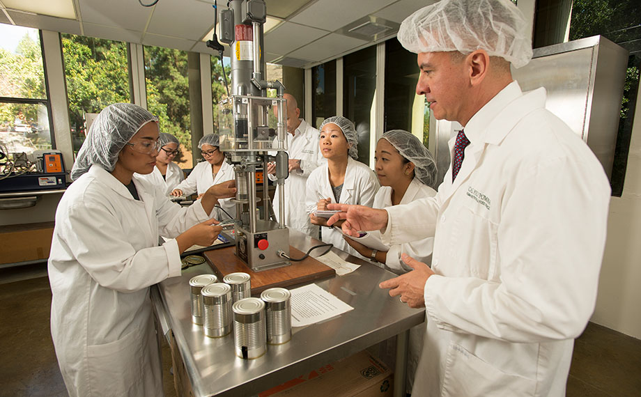 A professor and students, all clad in hairnets and lab coats, work in a kitchen lab.