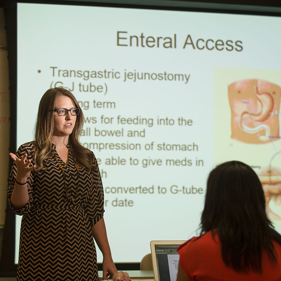 A woman lectures in front of a PowerPoint presentation