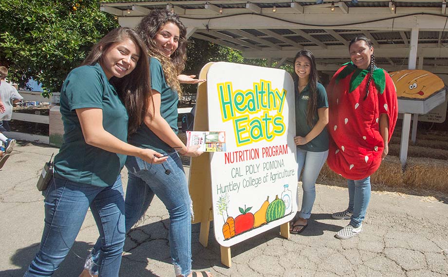 Four female nutrition students pose by a "Healthy Eats Nutrition Program" sign at the LA County Fair