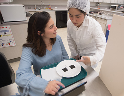 A student in lab coat gives another student food samples to test.