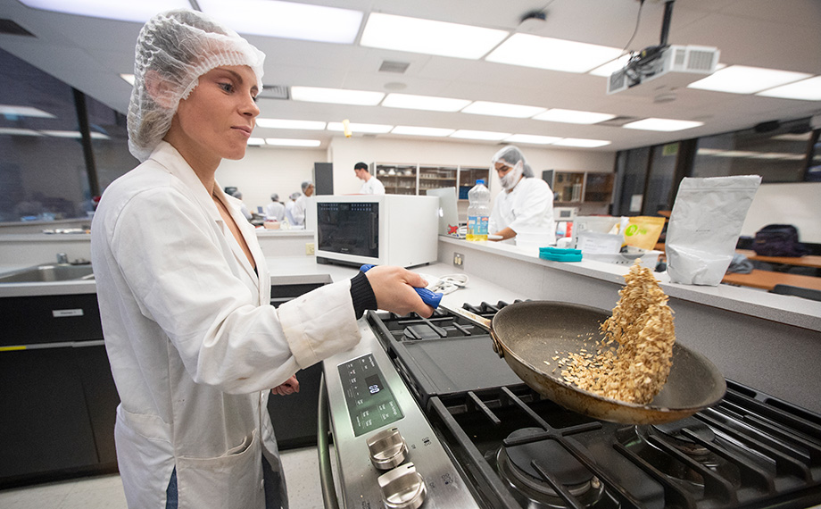 A female student in lab coat and hair net tosses food in a pan over a stove.