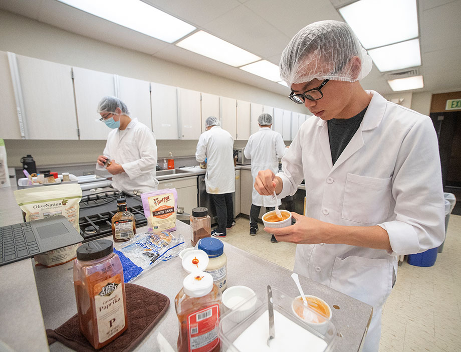 A student in hair net and lab coat works in a kitchen lab.