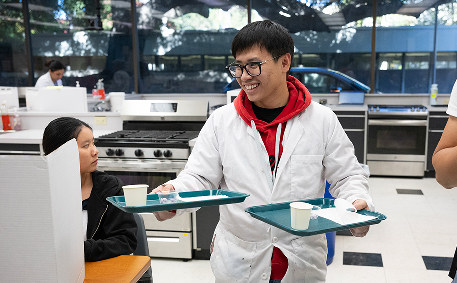A male student smiles and carries trays with food samples in a lab.