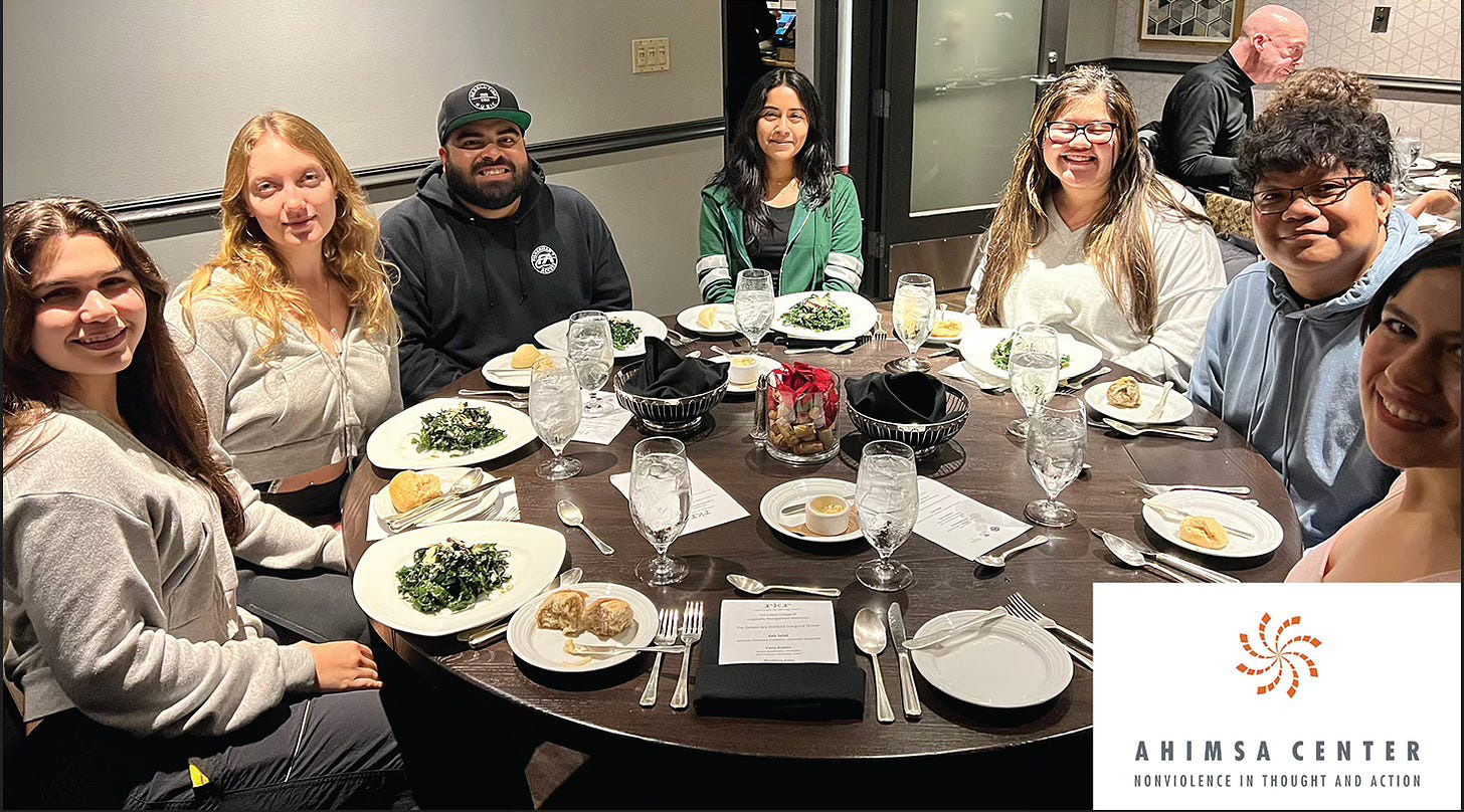 Group of Cal Poly Pomona students seated around a round dinner table, smiling and enjoying a formal meal at an event hosted by the Ahimsa Center, with the Center’s logo and tagline 'Nonviolence in Thought and Action' visible in the corner.