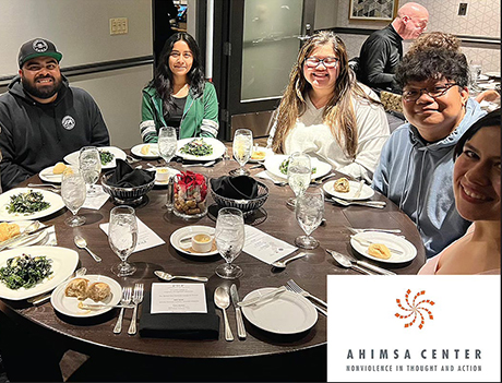 Group of Cal Poly Pomona students seated around a round dinner table, smiling and enjoying a formal meal at an event hosted by the Ahimsa Center, with the Center’s logo and tagline 'Nonviolence in Thought and Action' visible in the corner.