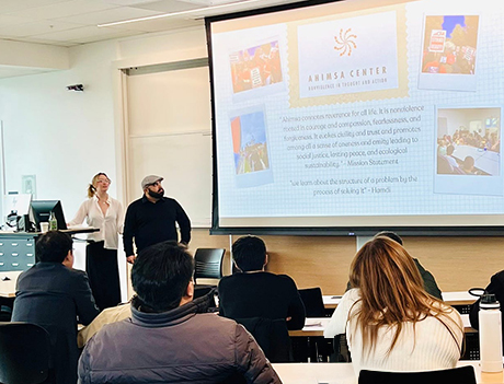 Two students present at the front of a classroom during the Oxford Consortium for Human Rights at Sonoma State University, with a slide displaying the Ahimsa Center and quotes about nonviolence projected behind them. Attendees are seated and listening.