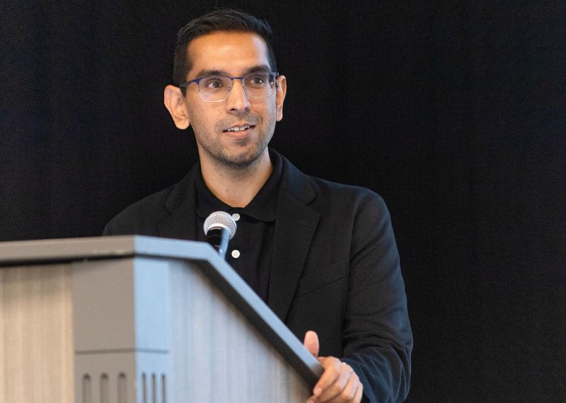 An alumnus in a black blazer speaks at a podium with a microphone during a formal presentation against a dark background.