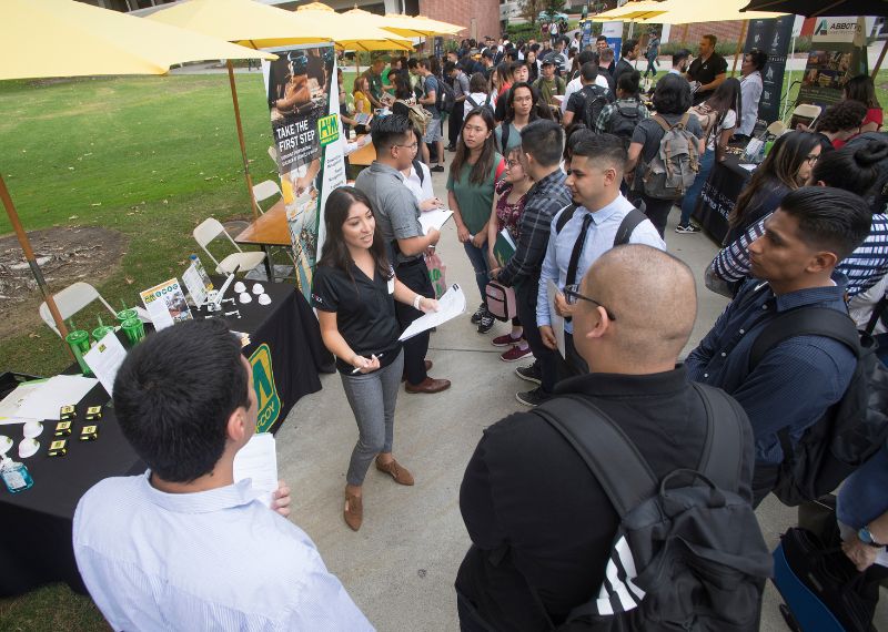 A recruiter speaks to a group of students at a bustling outdoor career fair with booths and informational banners.