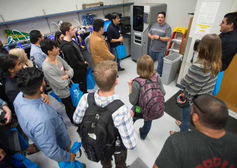 A group of students gathers around a speaker in a technology lab with 3D printers and advanced equipment.