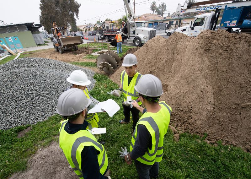 Construction workers in hard hats and safety vests review documents at a worksite with large equipment, gravel, and dirt piles