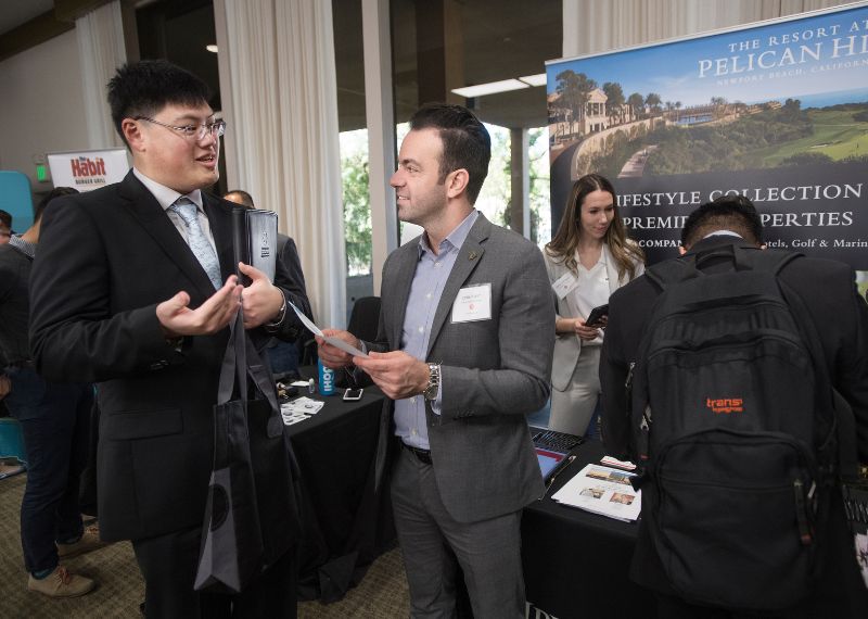 Two men in business attire have a conversation at a career fair booth promoting a luxury resort property.