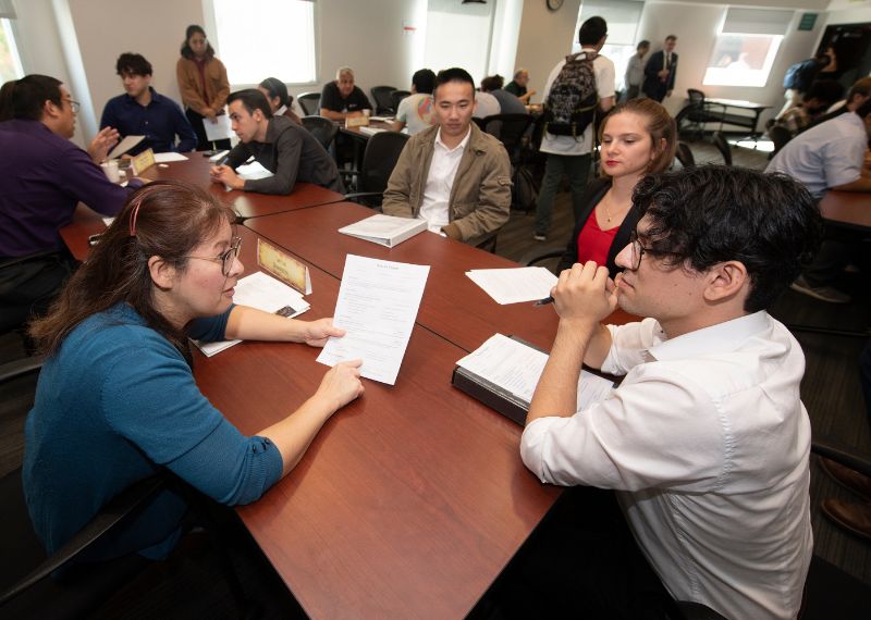 articipants engage in a resume review workshop, seated at tables and exchanging documents in a professional setting.