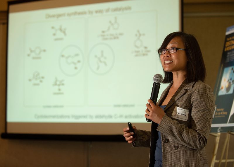  A presenter with a microphone explains a scientific diagram projected on a screen during a research presentation.