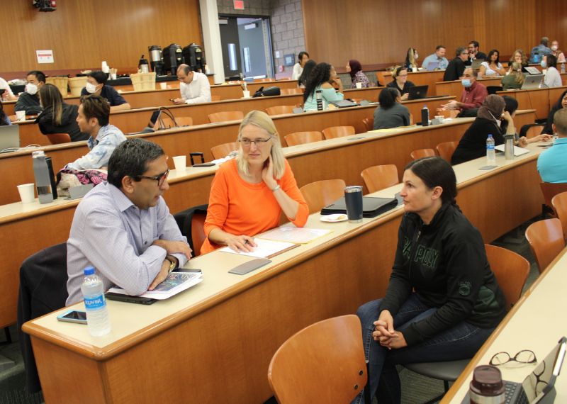 Three attendees in a lecture hall collaborate on a discussion during a workshop, surrounded by others engaged in group conversations.