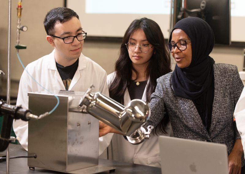 Three individuals in a laboratory setting, wearing lab coats and a headscarf, gathered around laboratory equipment with a laptop in the foreground.