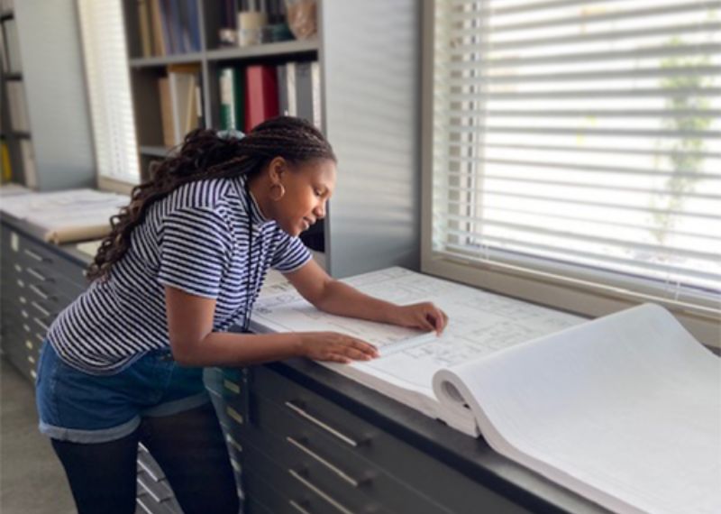 A woman in a striped shirt examines architectural blueprints in a well-lit office space.
