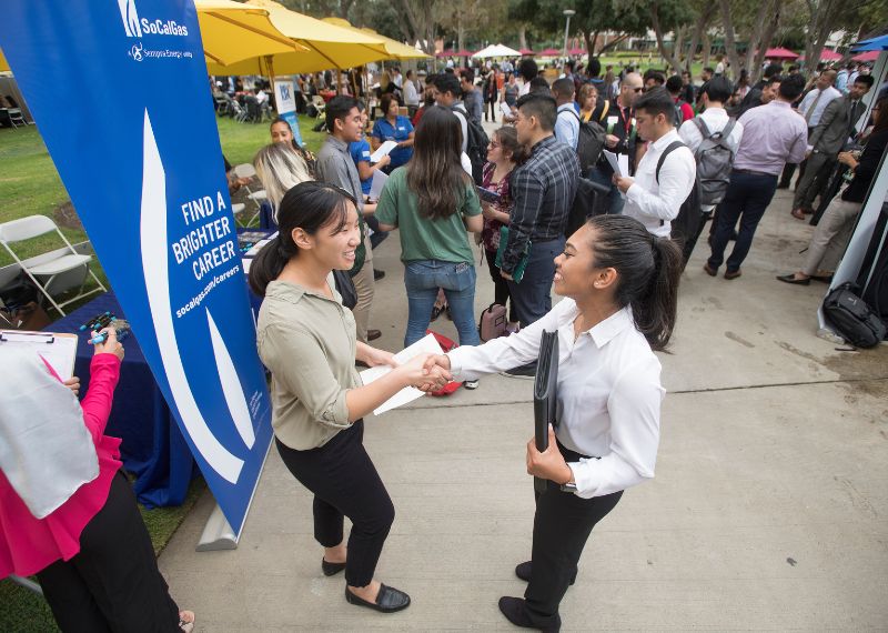 Two women shake hands at a busy outdoor career fair with informational booths in the background.