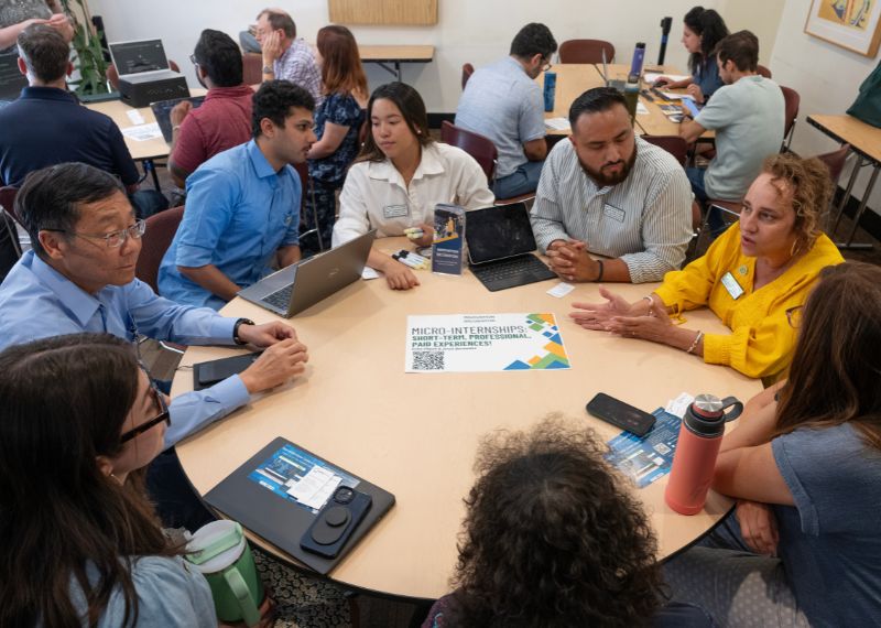 People sit around a table discussing micro-internship opportunities while using laptops and printed materials.