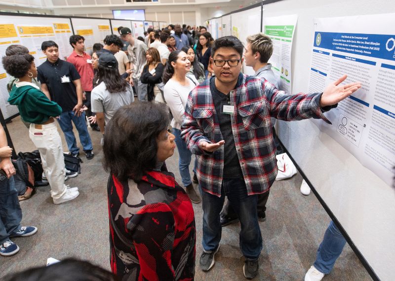 A student in a red plaid shirt explains a research poster to a woman at a busy academic showcase event.