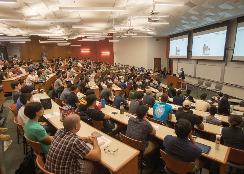 A large group of students listens attentively to a speaker during a presentation in a lecture hall.