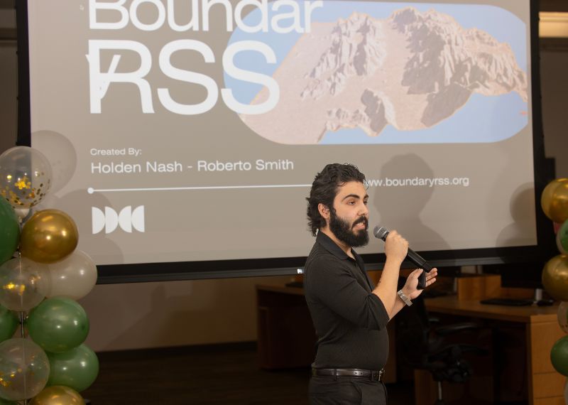  A man with a microphone presents his project titled "Boundary RSS" in front of a large projector screen during an event.