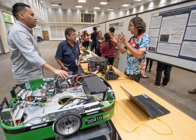 A student explains their robotics project to two attendees at a research showcase, with posters and robotic devices displayed on tables.