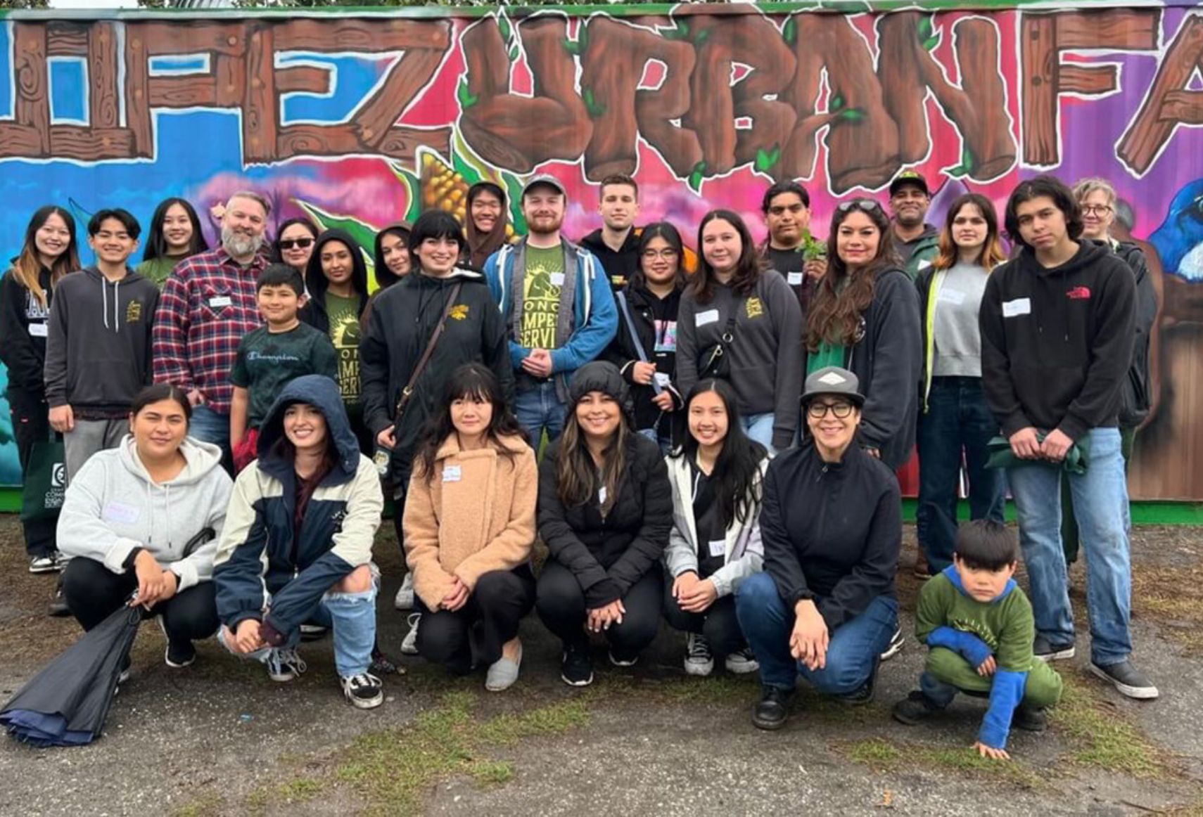 "A diverse group of individuals poses for a group photo in front of a mural that reads 'Urban Farm' during an outdoor event."