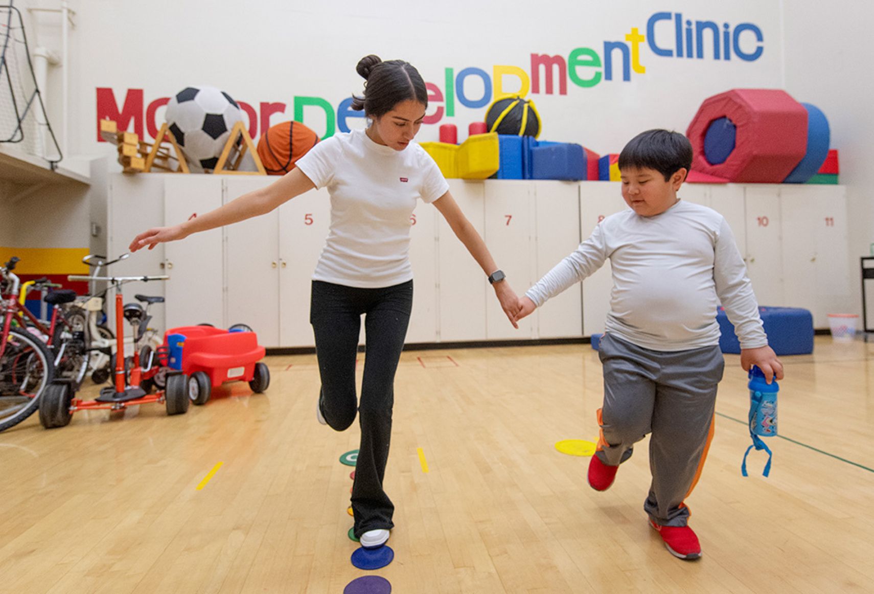 "A woman guides a young boy through a balance exercise in a colorful gym, indicating a developmental clinic."